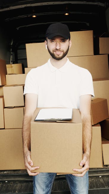 A man wearing a black cap, white polo shirt, and blue jeans is standing inside the back of a moving van, holding a medium-sized cardboard box with both hands. The van's interior is filled with neatly 